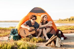 young couple camping during the summer and playing guitar while they drink coffee - sober summer vacation