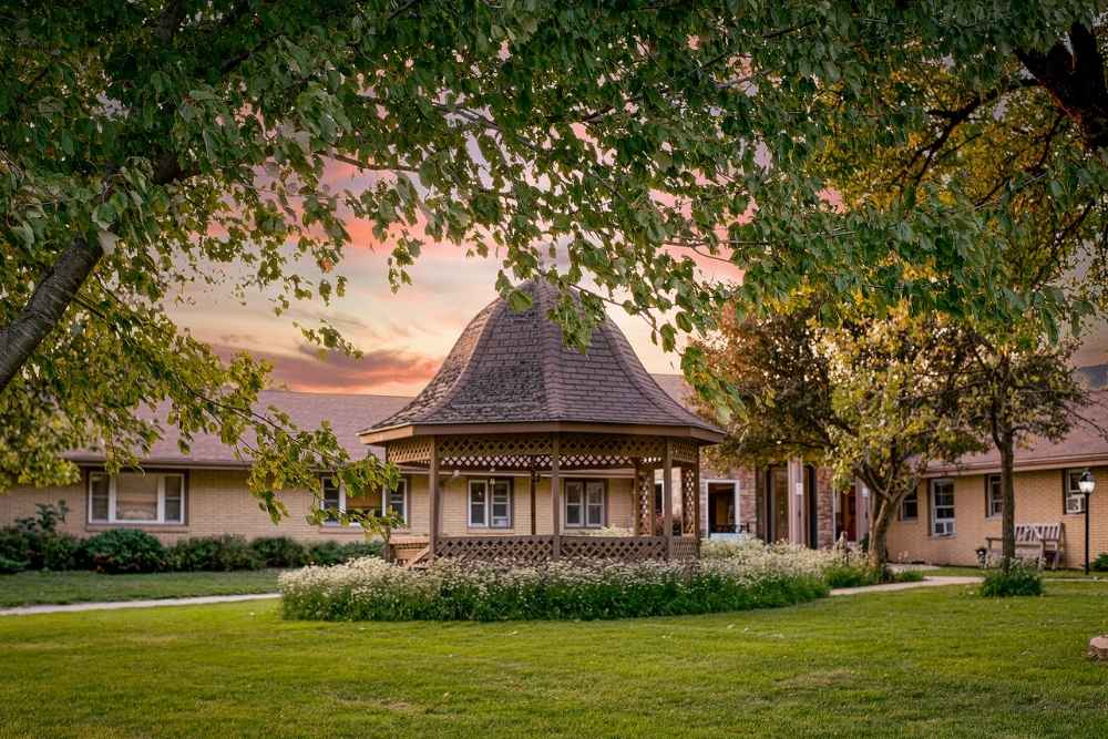 Peaceful courtyard with a central wooden gazebo surrounded by greenery and brick buildings, set against a colorful sunset sky.