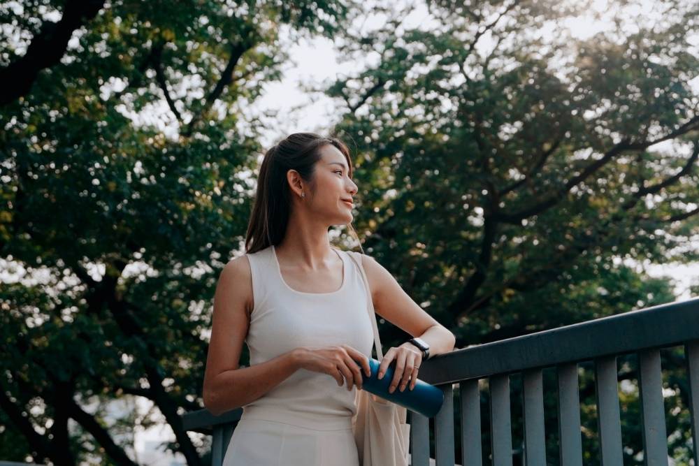 Woman standing outdoors on a bridge, holding a blue water bottle and looking thoughtfully into the distance, surrounded by trees and sunlight.