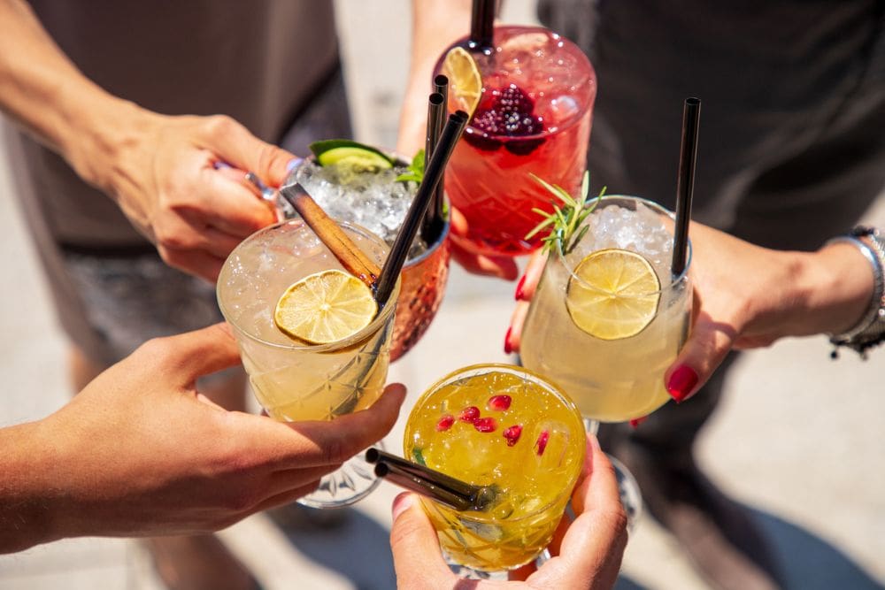 "Close-up of five people holding colorful mocktails garnished with fruits and herbs, toasting together outdoors in bright sunlight.