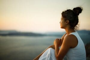 A woman sitting peacefully outdoors at sunset, gazing into the distance over a calm body of water, suggesting reflection, mindfulness, or healing.