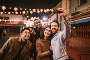 A group of five friends stands close together outdoors at night, smiling as one person takes a selfie. Decorative lanterns hang overhead, creating a festive atmosphere.