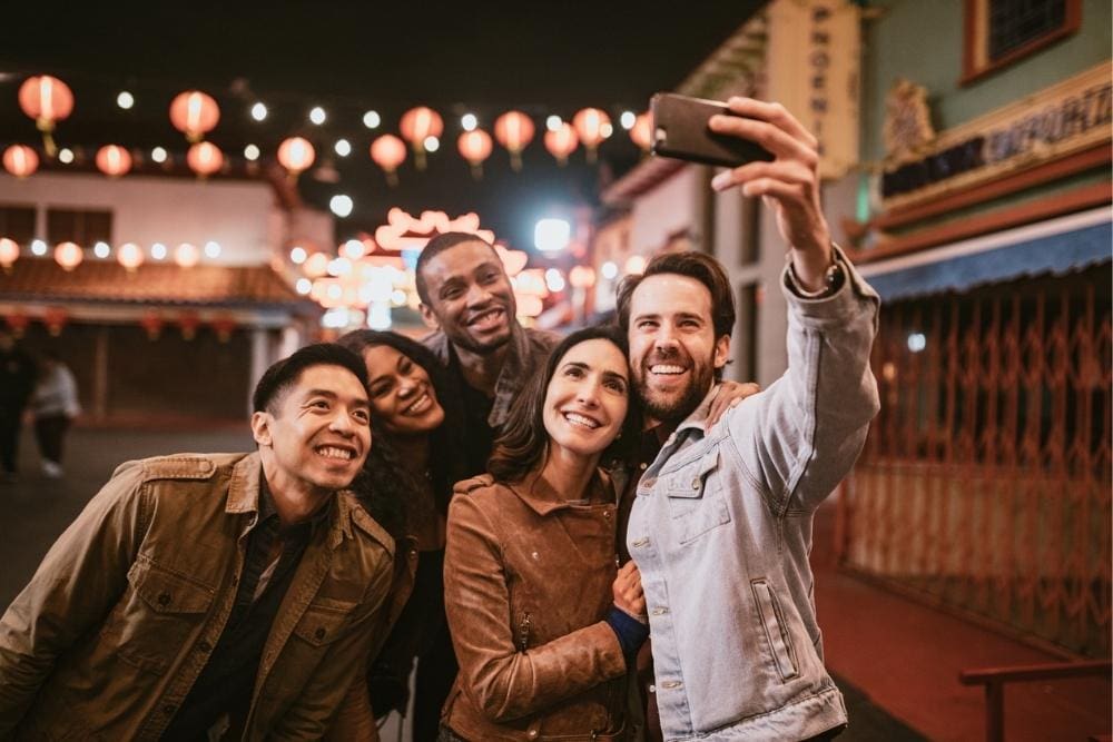 A group of five friends stands close together outdoors at night, smiling as one person takes a selfie. Decorative lanterns hang overhead, creating a festive atmosphere.