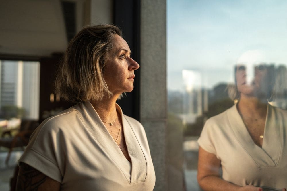 A middle-aged woman stands indoors, gazing thoughtfully out a large window. Her reflection is visible in the glass, and she appears pensive or reflective as natural light softly illuminates her face. Urban buildings and trees can be seen in the background.