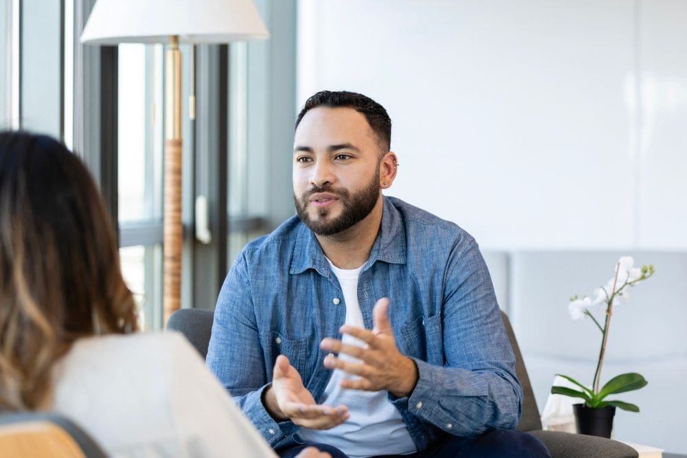 A man sits across from a woman in a bright, modern office setting, engaged in a thoughtful conversation. He gestures with his hands while speaking, suggesting active participation in a therapy or counseling session.