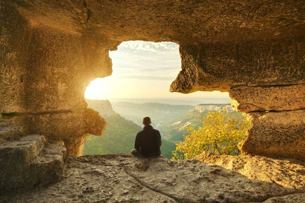 Person sitting on a rock ledge overlooking a scenic valley at sunrise or sunset.