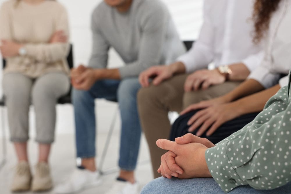Close-up of people sitting in a support group circle, with hands clasped or resting on laps, symbolizing recovery, connection, and shared experience.
