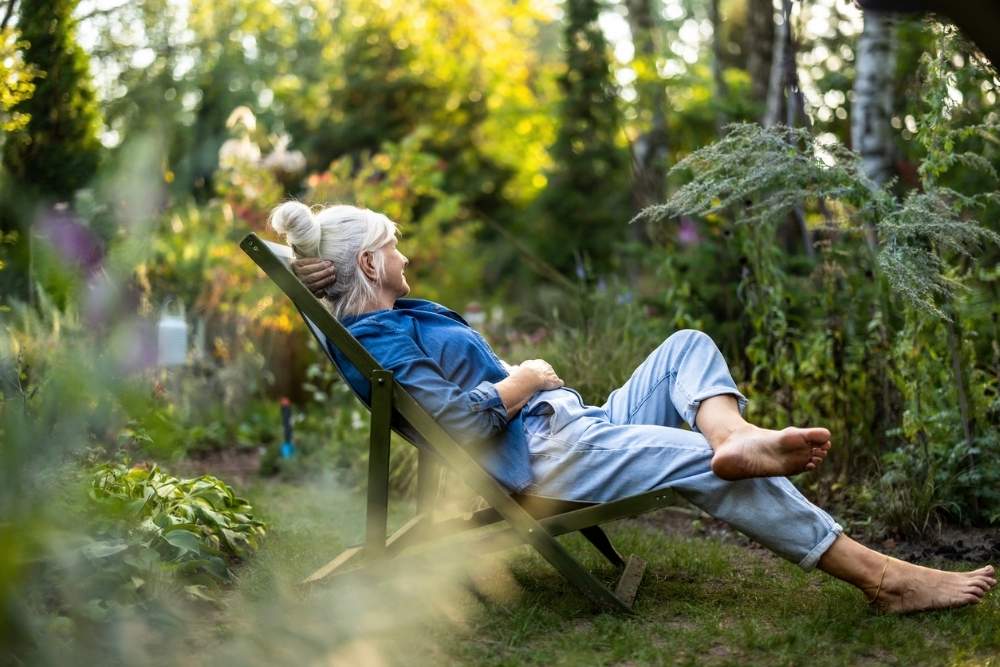 An older woman relaxes barefoot in a reclining chair in a lush garden, surrounded by greenery and sunlight, enjoying a peaceful moment outdoors.