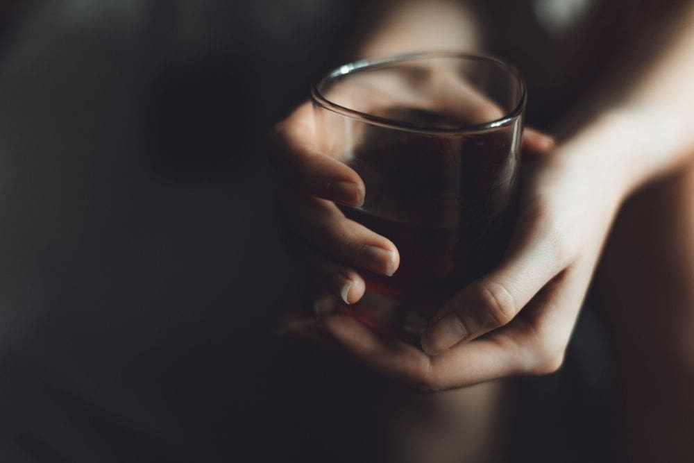 Close-up of hands holding a glass of dark alcohol in a dimly lit setting, symbolizing binge drinking and struggles with substance use.