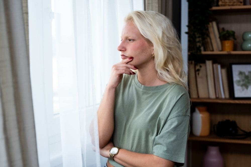 Young woman standing by a window with a thoughtful expression, resting her hand on her chin, symbolizing reflection and the need for support.