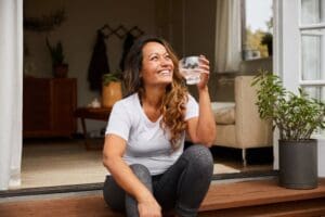 ature woman laughing while sitting alone on her patio steps outside drinking a glass of water