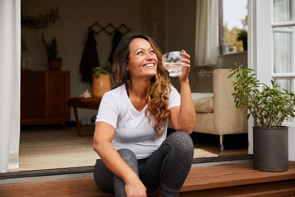 ature woman laughing while sitting alone on her patio steps outside drinking a glass of water
