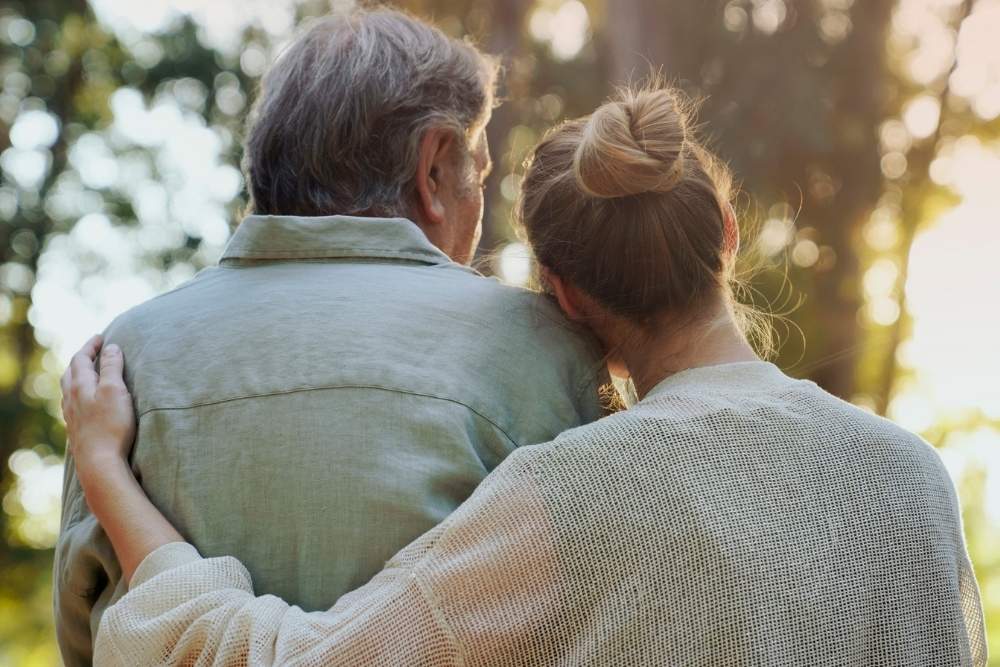 Woman resting her head on an older man’s shoulder while embracing him, standing together outdoors with sunlight filtering through trees.