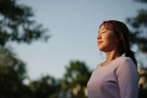 A woman stands outdoors with her eyes closed, taking a deep breath and enjoying the sunlight, symbolizing calm, balance, and mental well-being.
