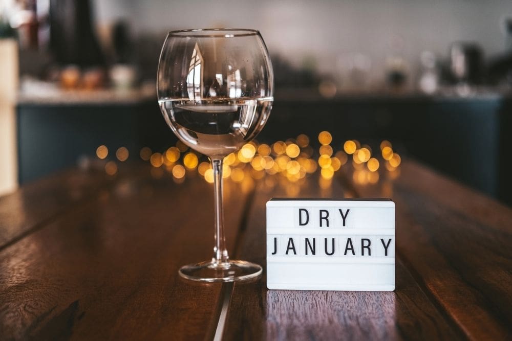 A glass of water on a table next to a lightbox sign reading ‘Dry January,’ with soft lights blurred in the background.