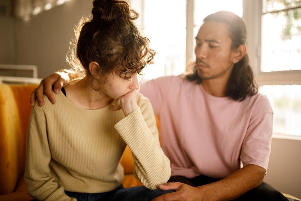 A man sitting beside a distressed woman, gently comforting her with a hand on her shoulder in a supportive, caring moment.