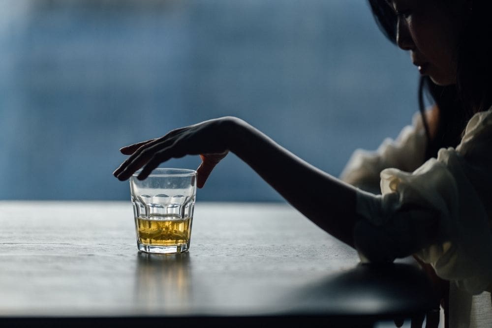 St. Gregory - Scientific Advances In AUD Treatment. A quarter filled glass of alcohol sits on a table. A shaddowy womans fingers are placed in the rim of the glass. The woman is in a dark room obscured by shadow.