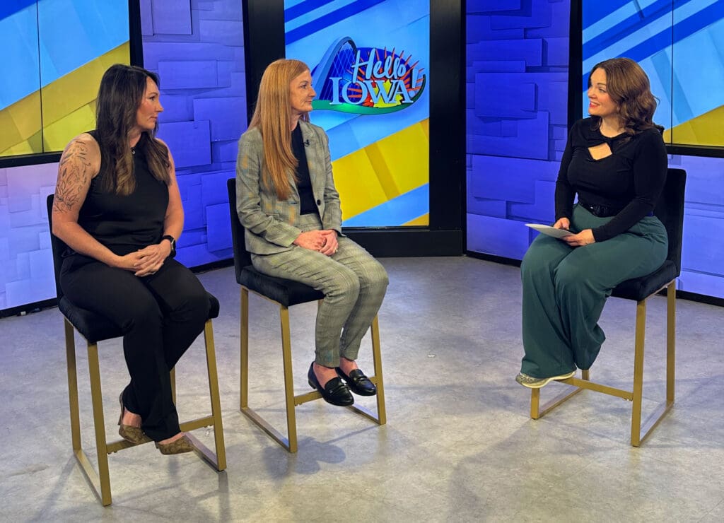Three women are seated on high seated chairs in a TV studio with "Hello Iowa" displayed in the background. They engage in a lively discussion.